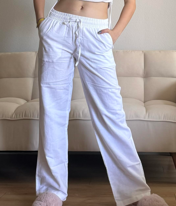 A person modeling casual white drawstring pants with their hands in their pockets, standing in front of a beige couch