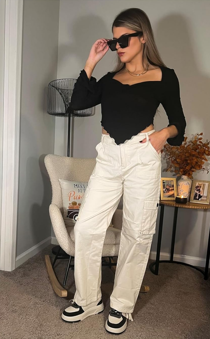 A person poses stylishly indoors, wearing a black top, white cargo pants, and sunglasses.