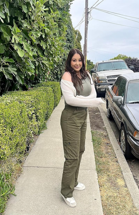 A person stands on a sidewalk, smiling at the camera while wearing cargo pants, a tank top, and a sheer jacket, with parked cars in the background.