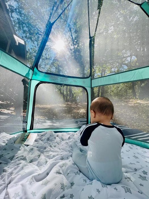 A baby wearing a long-sleeve onesie sitting in a mesh playpen outdoors