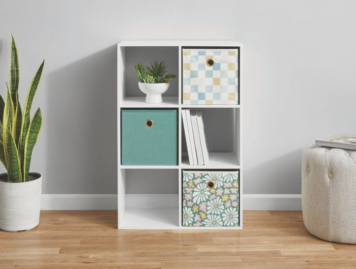 A white six-cube storage unit featuring colorful patterned bins, a potted plant on the floor, and a beige tufted ottoman nearby