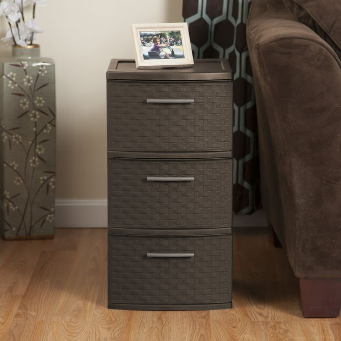 A three-drawer side table with a basket-weave texture, holding a small picture frame, placed beside a brown couch and a decorative floral vase in a cozy home setting.