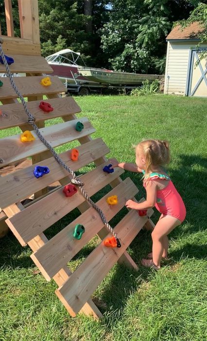 A young toddler in a swimsuit scaling a wooden play structure with colorful climbing grips in an outdoor setting