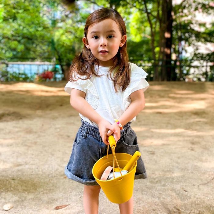 A young child standing in a park, holding a yellow beach bucket filled with toys.