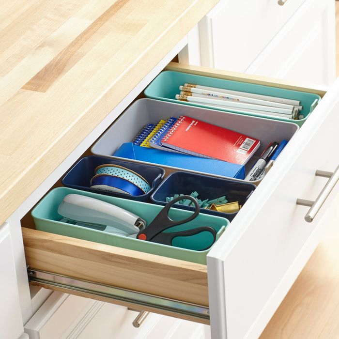An open drawer neatly organized with stationery items like notebooks, scissors, pens, pencils, tape, and paper clips all arranged in dedicated trays.