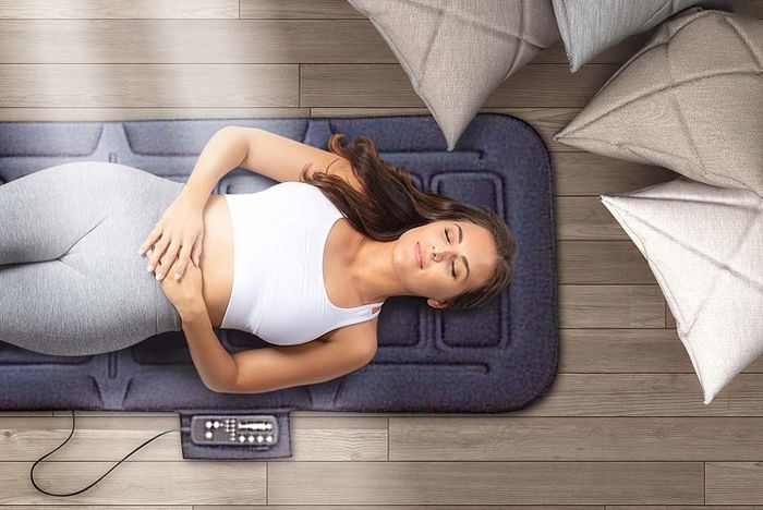 A person relaxing on a padded massage mat, which is slightly larger than a yoga mat and comes with a remote control for easy adjustments.