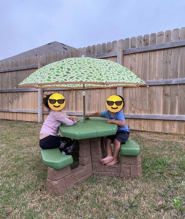 Two children sitting at a small outdoor play table with a patterned umbrella in a backyard, smiling.