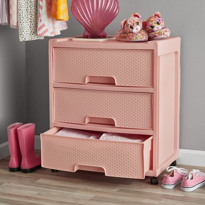 A pink storage dresser with three drawers, topped with a lamp and stuffed animal, while pink rain boots and slippers rest beside it