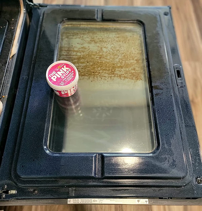 A reviewer’s photo showing a tub of The Pink Stuff on an oven door, with one side dirty and the other side sparkling clean