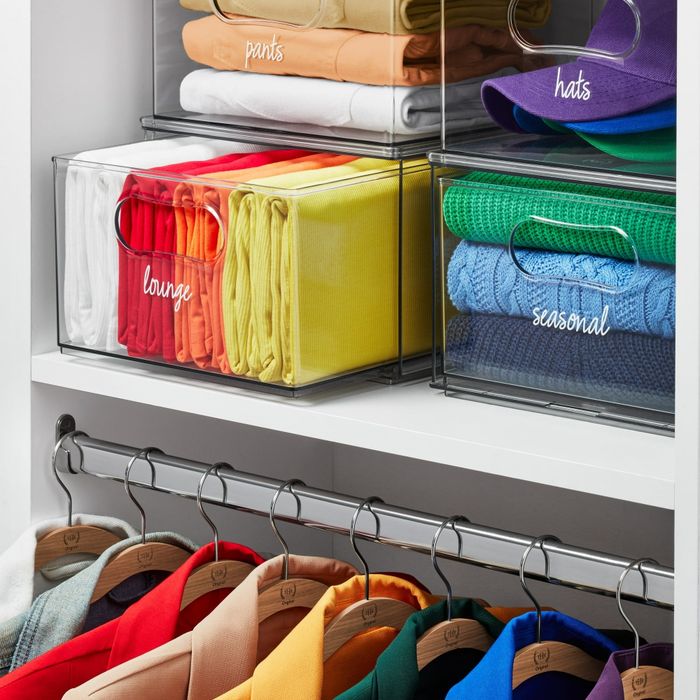 An organized closet with labeled clear bins for pants, hats, lounge, and seasonal items neatly placed on shelves, while colorful clothing hangs neatly below on hangers.