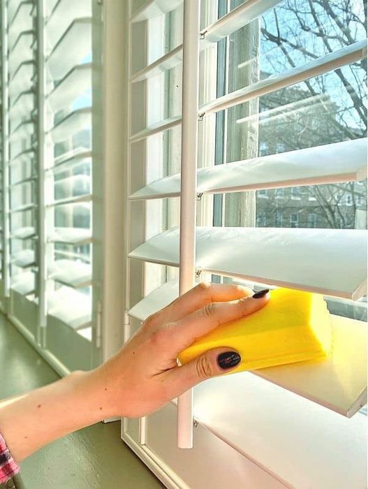 A hand using the yellow duster to wipe down window blinds