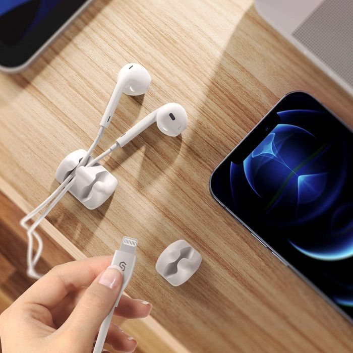 A person places a cable organizer on a wooden desk next to a pair of white earphones and a smartphone.