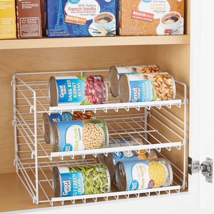 A kitchen cabinet with a wire shelf organizer holding canned goods such as beans, corn, and green beans, with boxes of Great Value coffee stacked above.