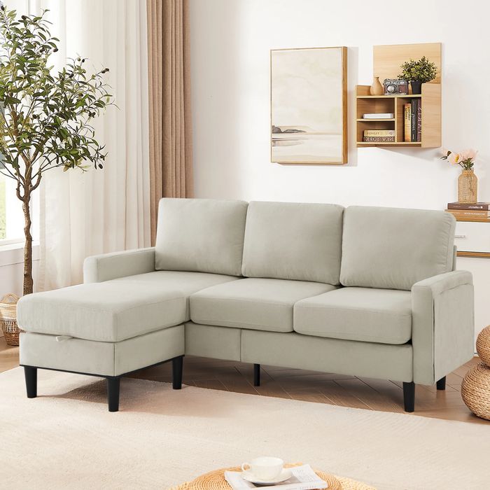 A beige sectional sofa with black legs placed in a bright living room, complemented by a wall painting, shelves, a potted plant, and a round woven rug