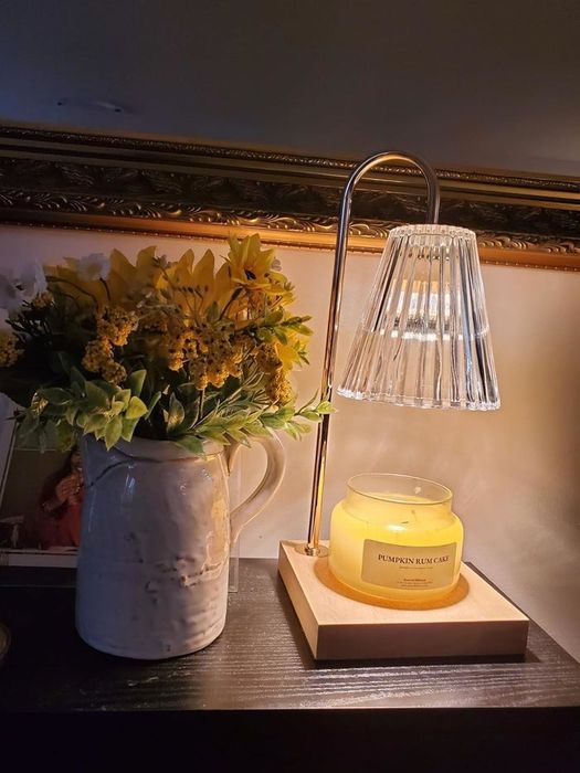 A tabletop candle warmer with a transparent shade lights up a pumpkin-scented candle, positioned beside a ceramic vase filled with sunflowers on a wooden surface