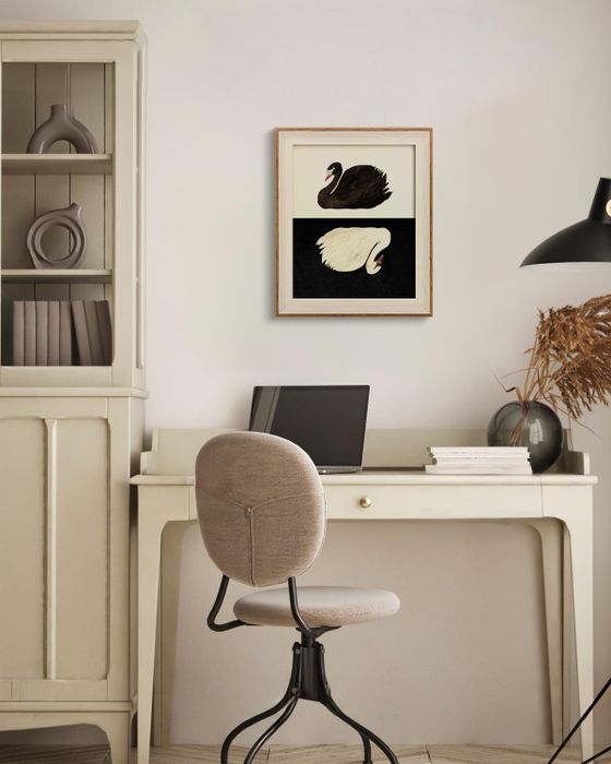 A home office setup featuring a beige desk and chair, a laptop, books, and a framed artwork depicting a black and a white swan hanging on the wall above.