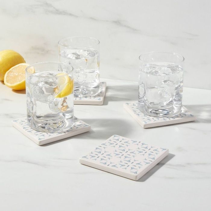 Three clear glasses filled with ice and topped with lemon slices rest on patterned coasters, accompanied by a fourth coaster and two lemons, all placed on a marble surface.