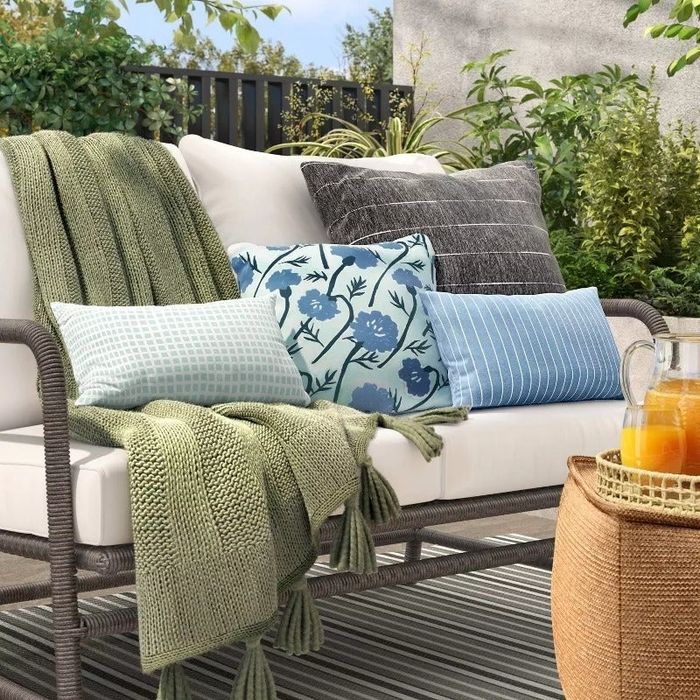 A cozy outdoor sofa with a green throw blanket, various patterned pillows, and a wicker basket on the side, holding a glass pitcher of fresh orange juice.