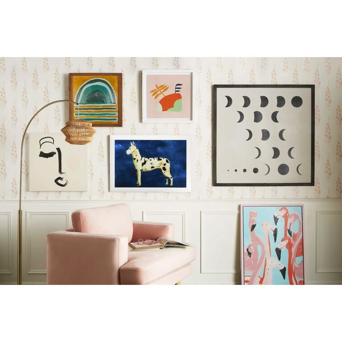 A chic living room wall featuring a variety of art pieces, with a pink chair completing the look. The art includes a vibrant rainbow, a majestic horse, lunar phases, abstract faces, and colorful flamingos.