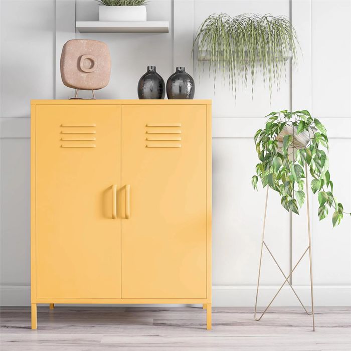 A yellow cabinet with dual handles, topped with decorative vases and a stone sculpture. Nearby, a tall potted plant on a stand and an indoor hanging plant add greenery to the space.