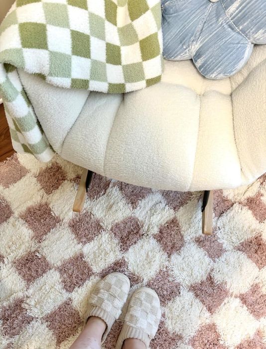 A cozy chair draped with a checkered blanket and cloud pillow, resting on a checkered rug; a person in checkered slippers stands nearby.