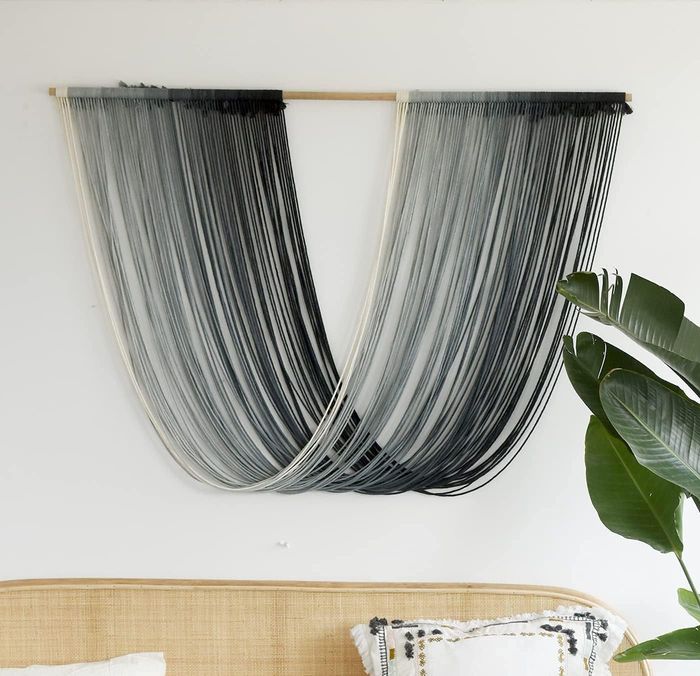 A modern yarn wall hanging displayed above a rattan bench with a cushion, next to a potted houseplant