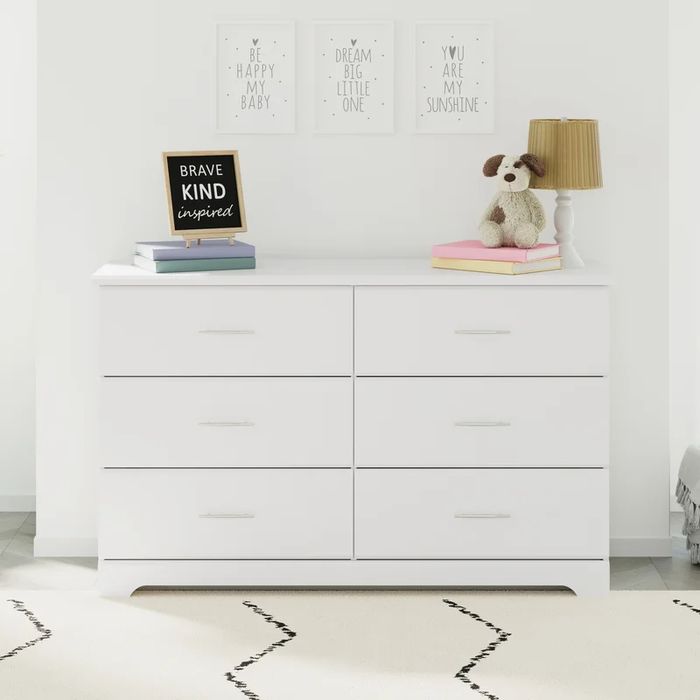 A white dresser with six drawers, topped with books, a lamp, and a plush dog.