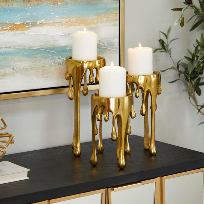Three pillar candles placed on golden candle holders with dripping accents, displayed on a table