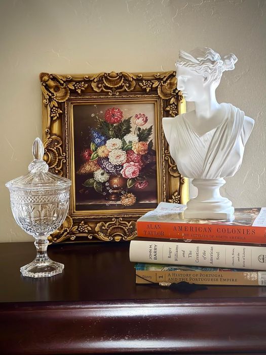 The sculpture is displayed on top of a pile of books, positioned next to a framed artwork.