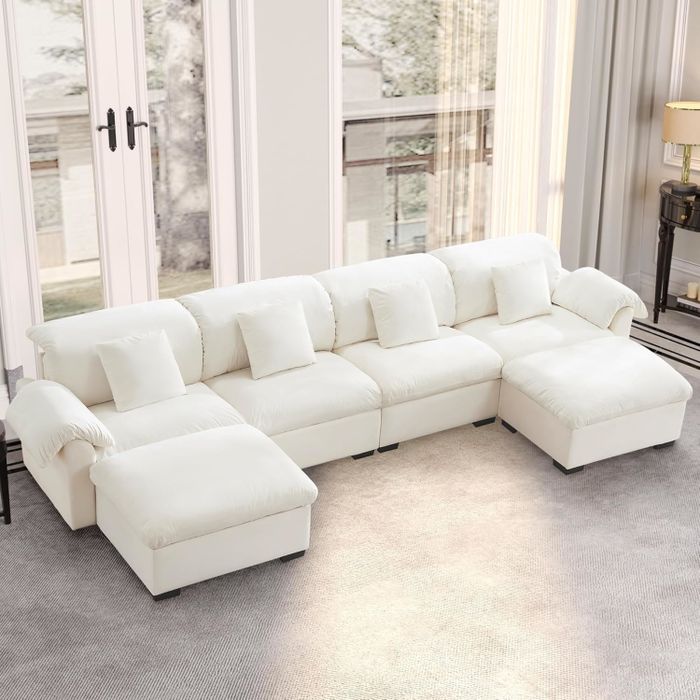 A spacious white sectional sofa adorned with multiple cushions, set in a bright, modern living room featuring large windows and a plush carpet.