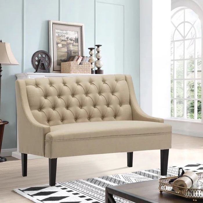 A beige tufted loveseat with black legs is placed in a bright and airy room, complete with a large window, a stylish lamp, framed artwork, and decorative accents in the background