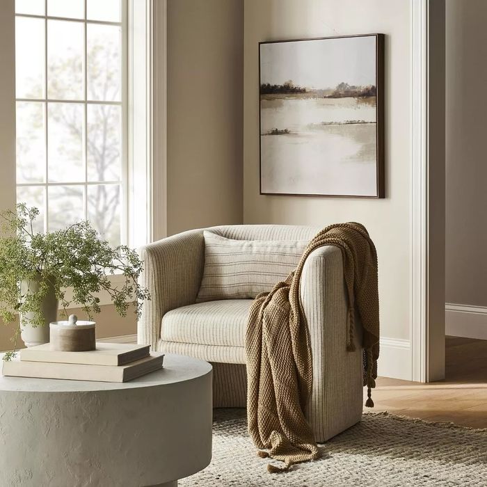 A cozy living room featuring a striped armchair draped in a knitted throw, a round table adorned with decor, and a landscape painting hanging on the wall.