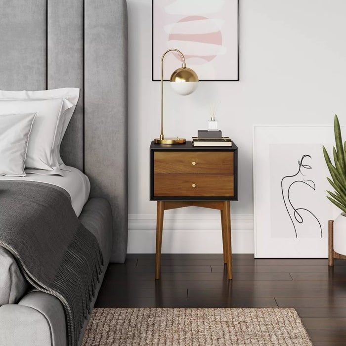 Modern bedroom featuring a gray upholstered bed, a wooden nightstand with a gold lamp, abstract wall art, and a potted plant