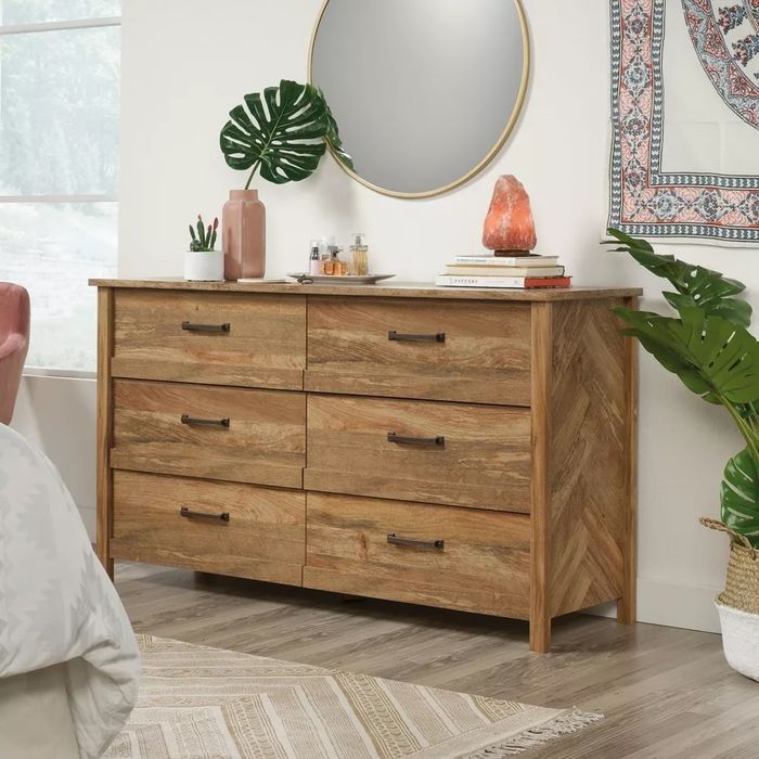 A wooden dresser with six drawers, potted plants, a round mirror, and decorative accents, nestled in a modern bedroom design.