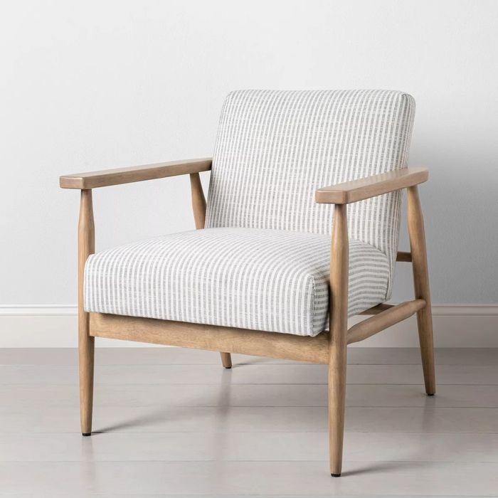 A modern armchair featuring light striped upholstery, wooden arms, and legs. It is placed on a light-colored floor, set against a simple, plain wall.