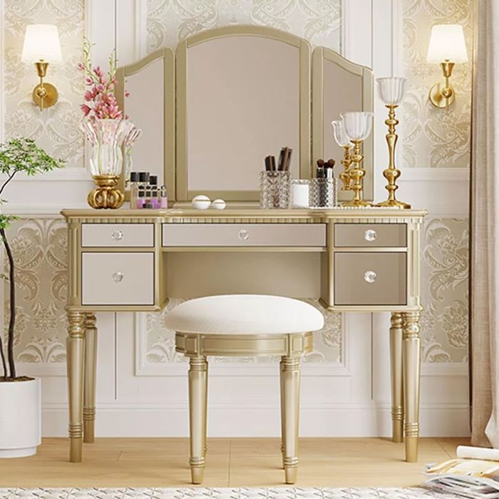 A luxurious vanity set complete with drawers, a mirror, and makeup items on top. The set also comes with a cushioned stool and is surrounded by elegant decor.