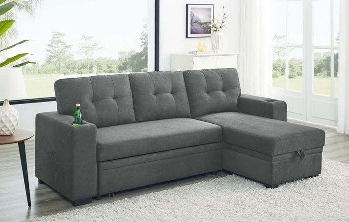 A modern gray sectional sofa with tufted cushions and a built-in cupholder armrest, placed in a bright, airy living room with large windows and a white rug