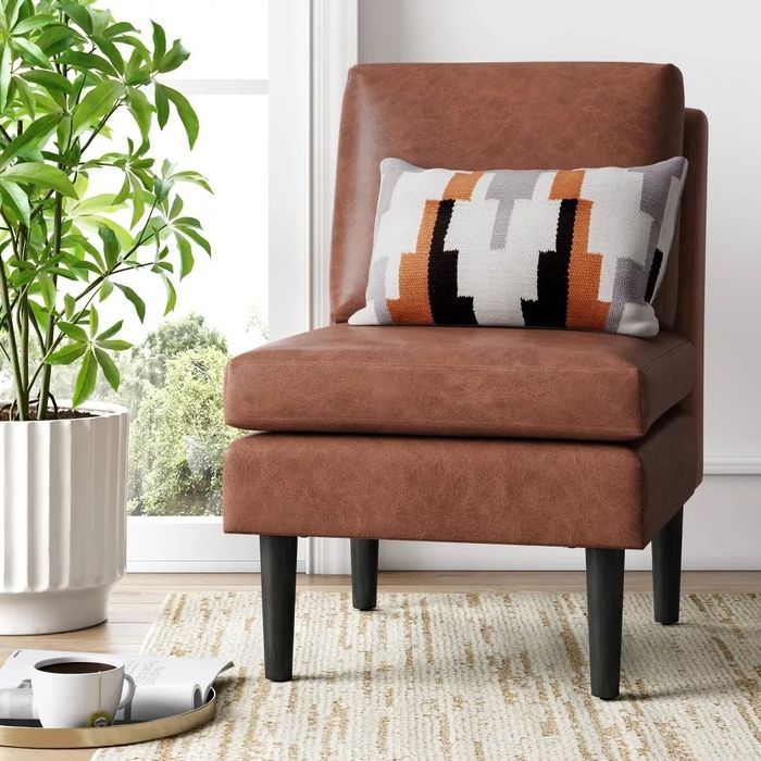 A brown leather accent chair with a stylish patterned cushion, placed next to a white pot containing a green plant. The setting features a bright and inviting living room atmosphere.