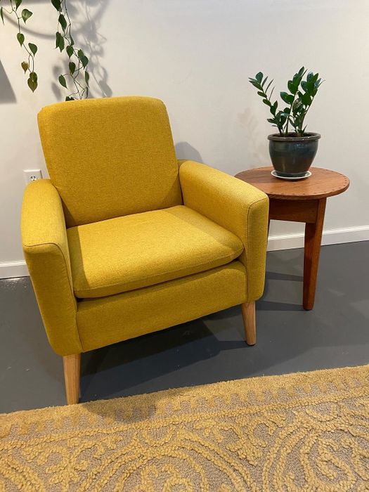 A mustard yellow armchair placed next to a small wooden table with a potted plant, atop a textured rug. It's perfect for creating a warm and inviting atmosphere in any room.