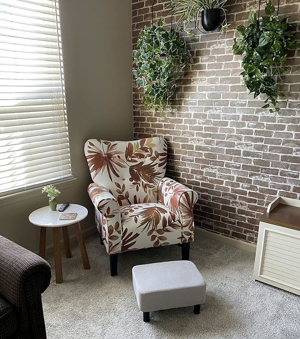 A cozy reading nook with a floral-patterned armchair, matching ottoman, small table, and hanging plants next to a brick wall.
