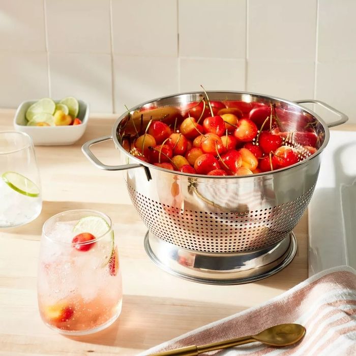 A stainless steel colander filled with cherries, sitting on a kitchen counter beside a cocktail garnished with fruit