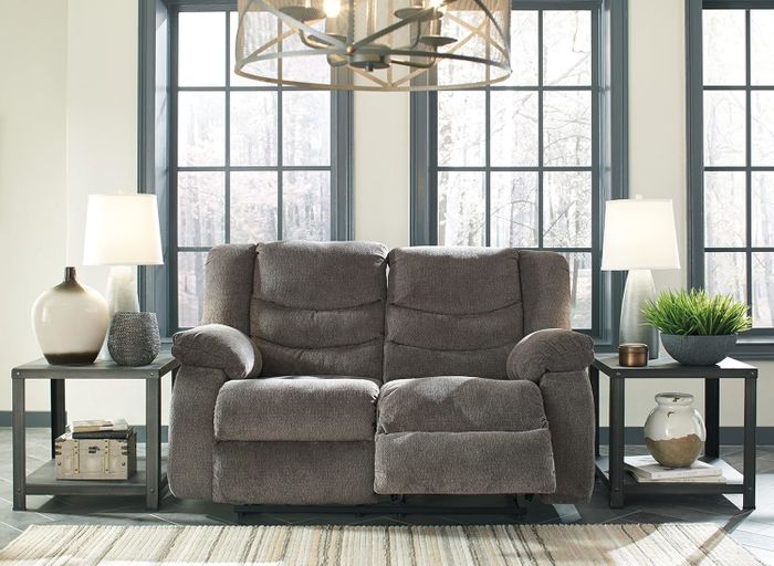 A cozy grey loveseat recliner nestled between two side tables with matching lamps and decorative accents, brightened by large windows in a spacious living room.