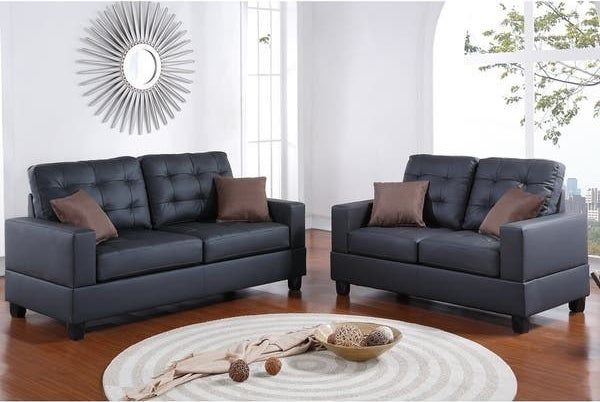 Product image of two sleek black faux leather couches with brown pillows in front of a round coffee table