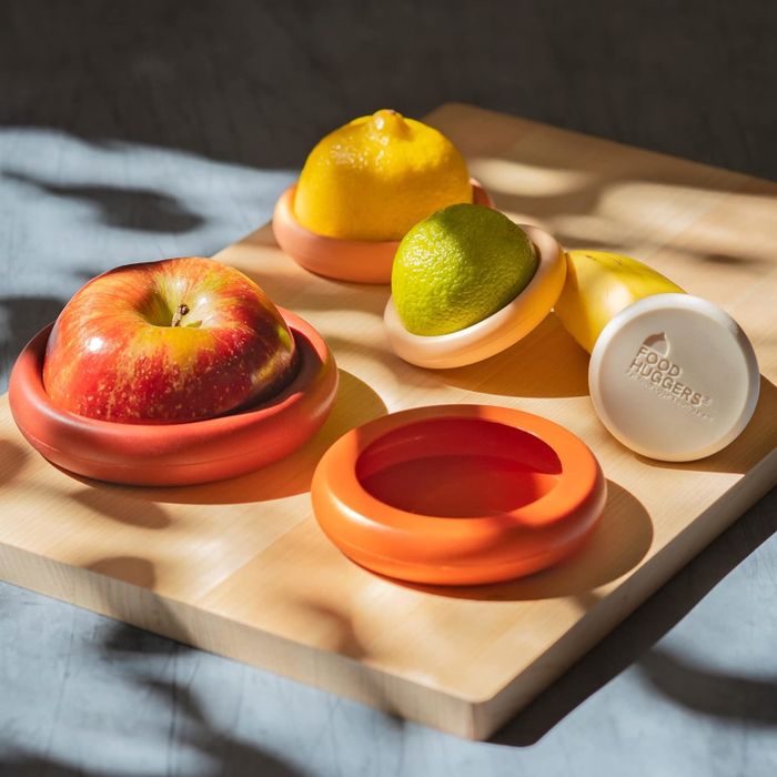 A partially sliced apple, lemon, and lime wrapped in silicone Food Huggers on a wooden cutting board, with an unused Food Hugger beside them