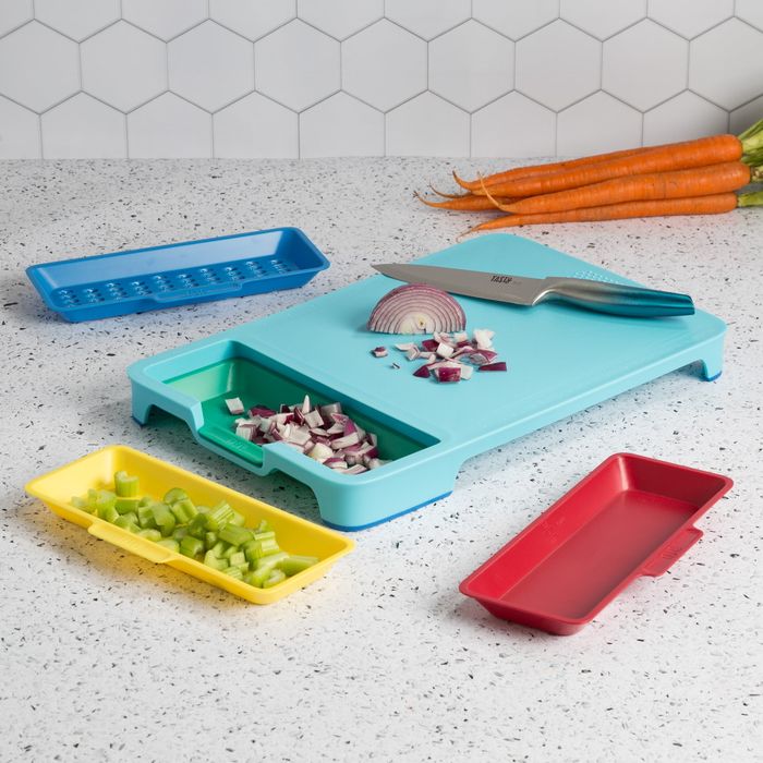 A cutting board with multiple compartments, a knife, diced onions, and celery neatly placed on a speckled countertop, with carrots visible in the background.