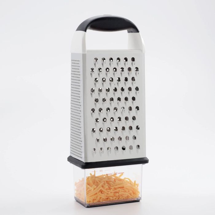 A metal box grater with a black handle positioned over a transparent container filled with freshly shredded cheese