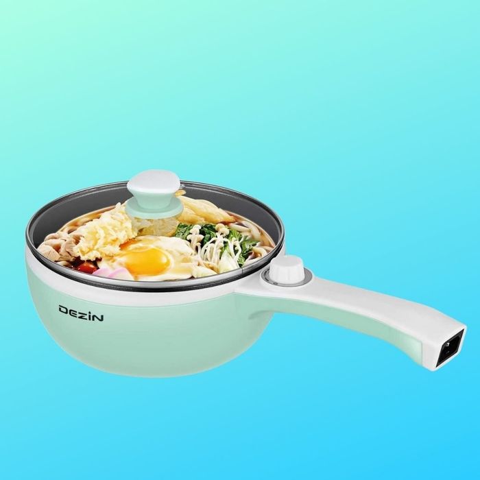 A white Dezin electric saucepan filled with an assortment of ingredients like noodles, veggies, and eggs, against a plain backdrop.