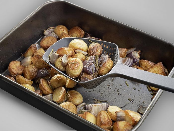 Roasted potatoes and onions in a baking tray with a slotted spoon lifting some of the pieces.