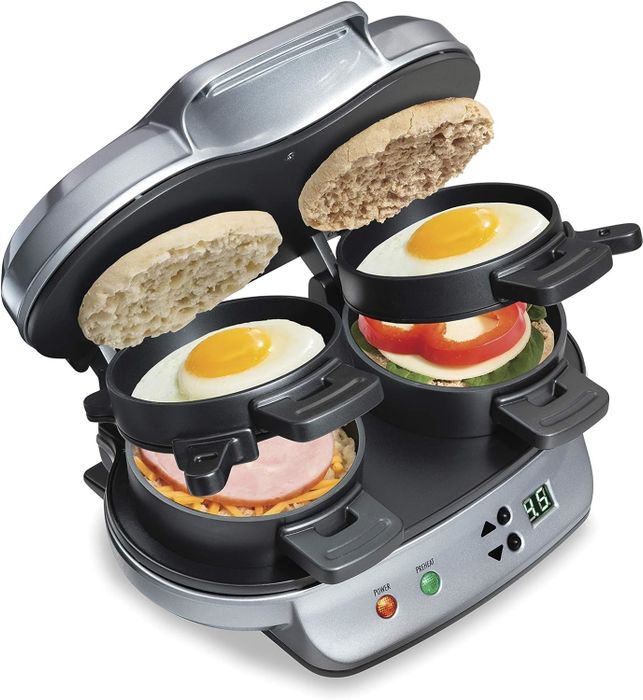 A sandwich maker preparing breakfast sandwiches with eggs, English muffins, ham, cheese, tomato, and spinach, partially open to show the cooking process
