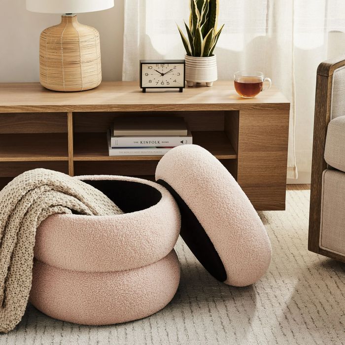 A blush-colored storage ottoman, with a cozy throw blanket tucked inside, sitting atop a white striped rug in a living room next to a wooden sideboard featuring a table lamp, a small clock, and a potted plant.